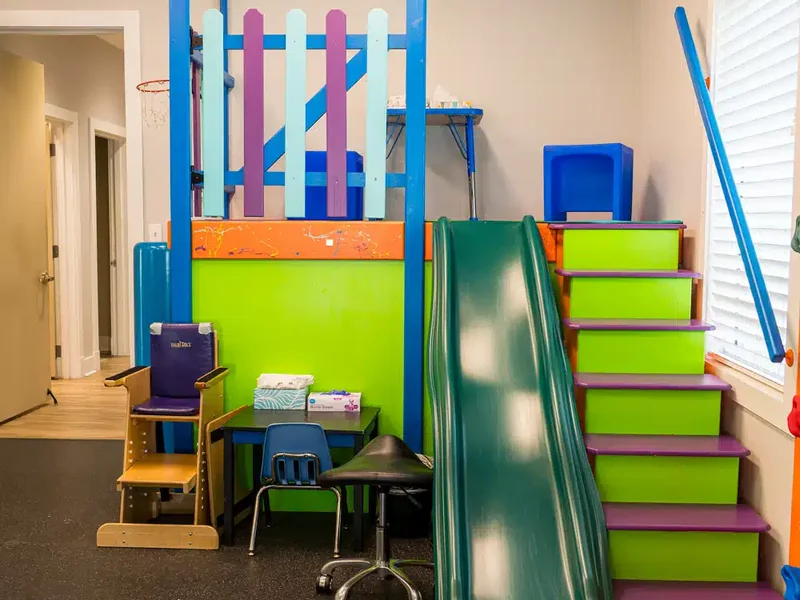 Therapy gym with a green slide, painted steps, a small table and a child sized chair